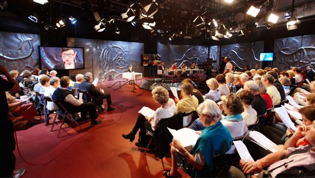 Zuschauer sitzen in einem Fernsehstudio vor einer Leinwand mit einer Frau.