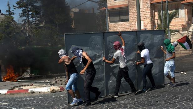Vermummte Männer schieben eine Metallbarriere vor einem brennenden Barrikade her.