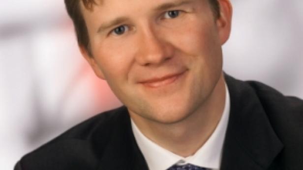 Ein Mann mit blonden Haaren, Anzug und Krawatte lächelt in die Kamera.