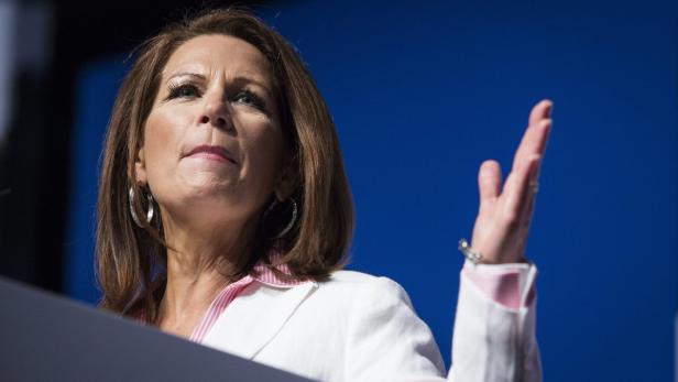 Eine Frau in einem weißen Blazer gestikuliert vor einem blauen Hintergrund.