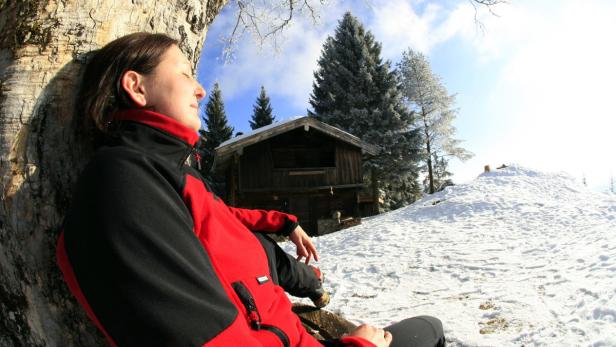 Eine Frau sitzt an einem Baum in einer verschneiten Winterlandschaft mit einer Hütte im Hintergrund.
