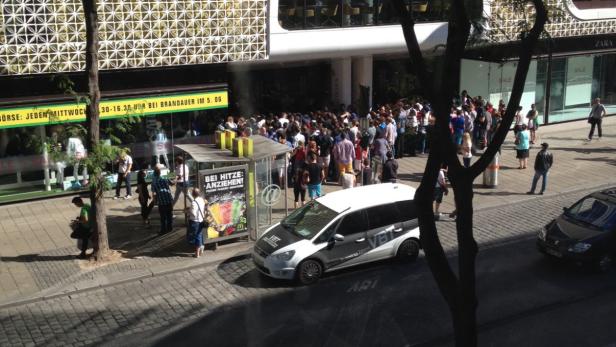 Eine lange Menschenschlange wartet vor einem Gebäude in der Sonne.