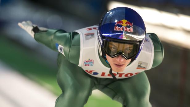 Ein Skispringer in grüner Montur und blauem Helm mit Red Bull-Logo in der Luft.