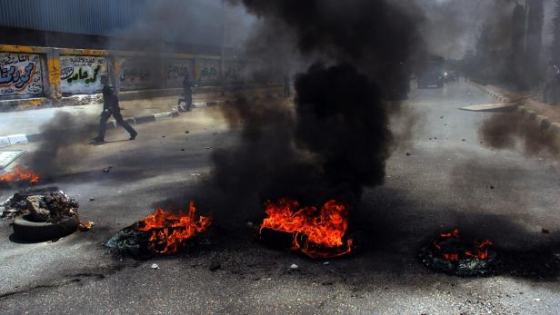 Auf einer Straße brennen mehrere Feuer, während im Hintergrund Personen zu sehen sind.