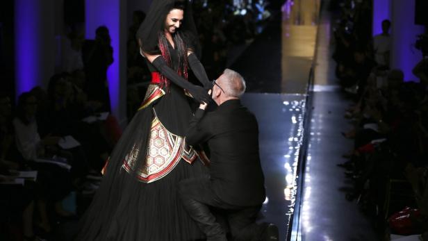 Ein Designer kniet vor einem Model in einem schwarzen Kleid auf dem Laufsteg.