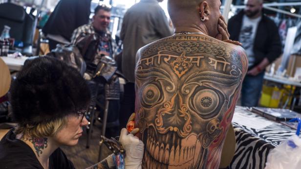 Eine Tätowiererin sticht ein großes Tattoo mit einem Totenkopf auf den Rücken eines Mannes.