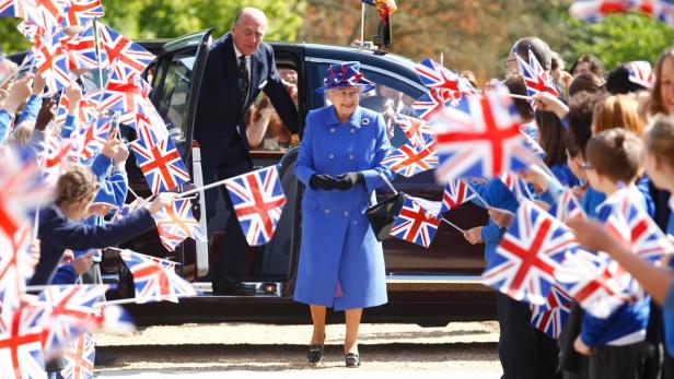 Königin Elisabeth II. steigt aus einem Auto, umgeben von Kindern mit Union Jack-Flaggen.