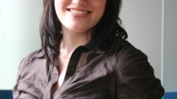 Eine lächelnde Frau mit Brille und dunklem Haar trägt eine braune Bluse mit einer Halskette.