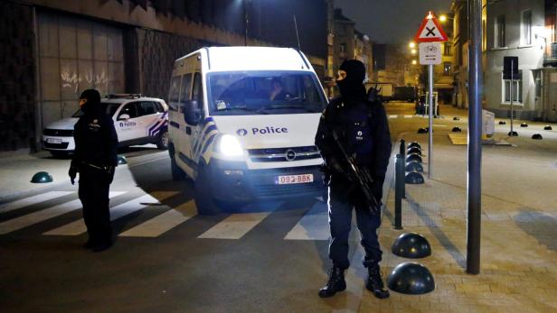 Ein Polizist mit Sturmhaube und Gewehr steht vor einem Polizeiwagen in einer nächtlichen Straßenszene.