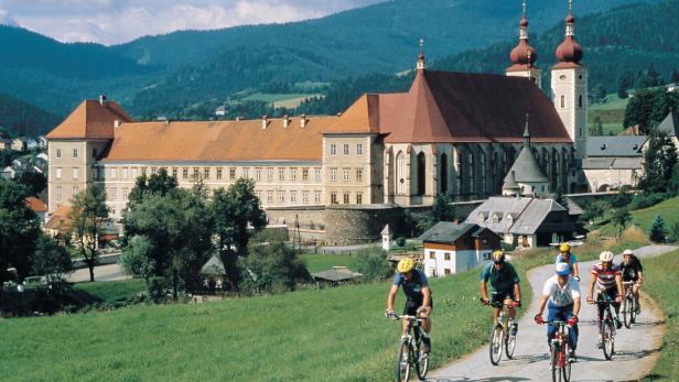 Eine Gruppe Radfahrer fährt auf einer Straße vor dem Stift Neuburg in Österreich.