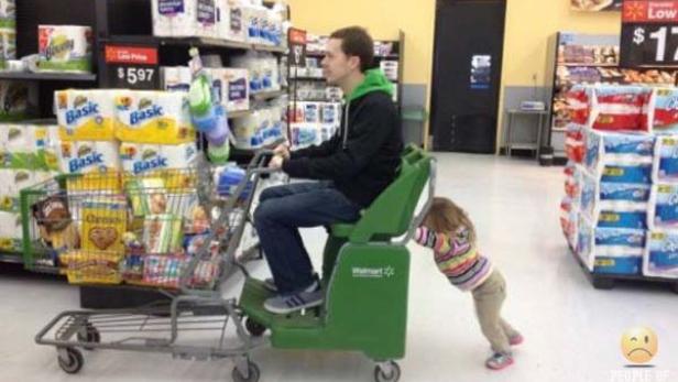 Ein Mann fährt in einem Walmart auf einem motorisierten Einkaufswagen, während ein kleines Kind ihn schiebt.