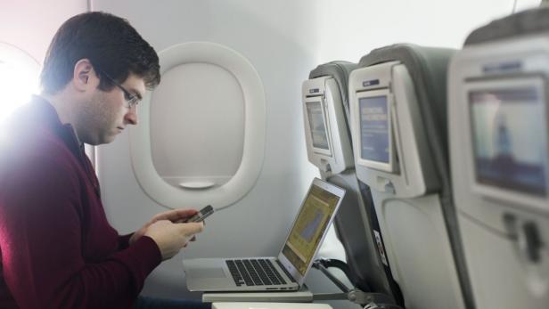 Ein Mann sitzt im Flugzeug, tippt auf seinem Handy und hat einen Laptop vor sich.