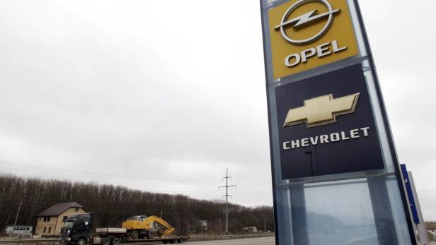 Ein Schild mit den Logos von Opel und Chevrolet vor einer trüben Landschaft.