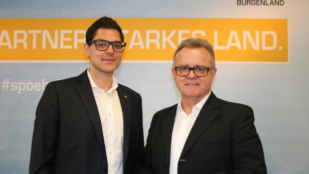 Zwei Männer in Anzügen posieren vor einem Hintergrund mit der Aufschrift „Partner. Starkes Land“.