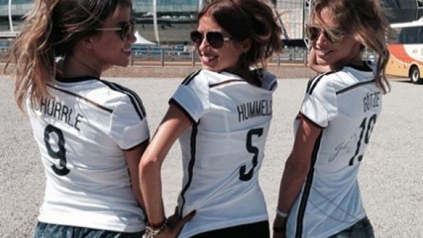 Drei junge Frauen in Deutschland-Trikots posieren vor einem Fußballstadion.