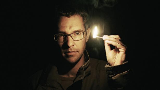 Ein Mann mit Brille hält ein brennendes Streichholz in der Hand.