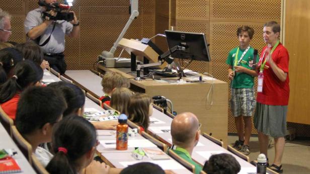 Ein Vortragssaal mit Kindern, einem Kameramann und zwei Personen vor einem Tisch mit einem Monitor.