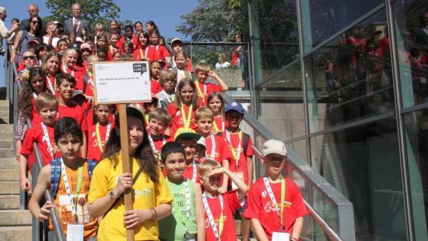 Eine Gruppe Kinder steht auf einer Treppe und hält ein Schild mit der Aufschrift „160: Welche Sprachen spricht die Stadt?“.