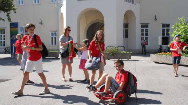Eine Gruppe von Kindern und eine Frau sind vor einem Gebäude unterwegs, ein Junge sitzt auf einem Pedal-Gokart.