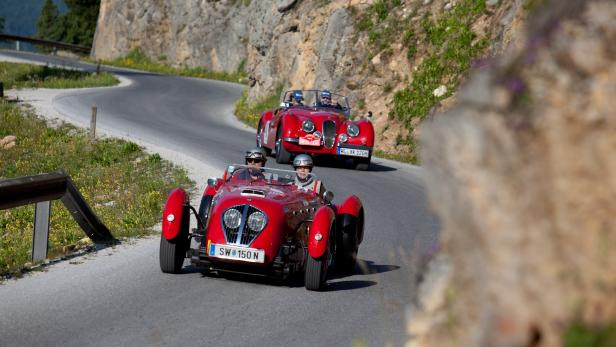 Zwei rote Oldtimer fahren auf einer kurvenreichen Bergstraße.