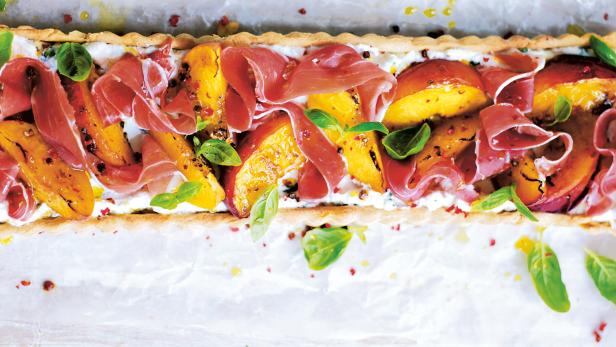 Eine rechteckige Tarte mit Ricotta, gegrillten Pfirsichen, Prosciutto und Basilikum belegt.