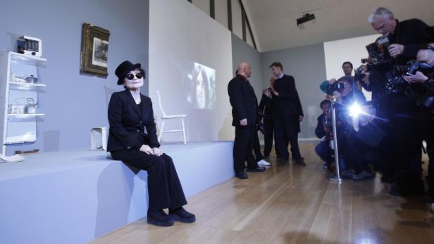 Yoko Ono sitzt vor einer Installation, während sie von Fotografen umgeben ist.
