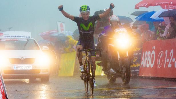 Ein Radrennfahrer überquert im Regen mit erhobenen Armen die Ziellinie.