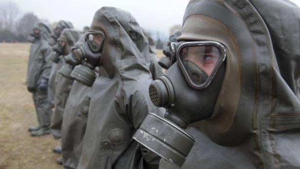 Eine Reihe von Personen in Schutzanzügen und Gasmasken steht im Freien.