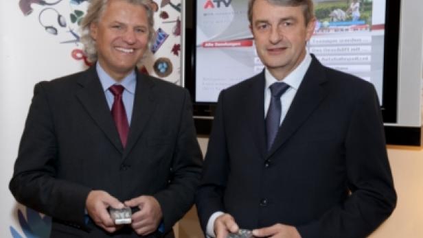 Zwei Männer in Anzügen halten Fernbedienungen vor einem ATV-Fernseher.