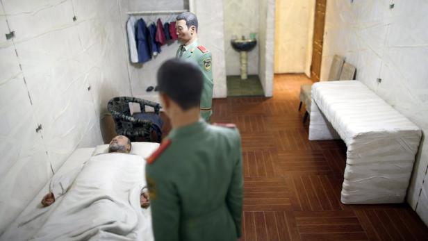 Ein Mann liegt in einem kargen Raum auf einem Bett, während zwei Soldaten ihn bewachen.