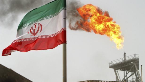 Die iranische Flagge weht vor einer Erdölraffinerie, aus der Flammen schlagen.