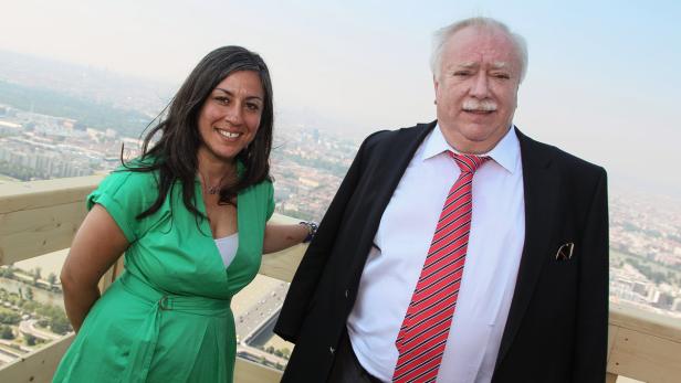 Eine Frau in einem grünen Kleid und ein Mann mit Anzug stehen vor einer Stadtlandschaft.