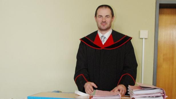 Ein Mann in einer Robe steht hinter einem Schreibtisch mit Aktenordnern.