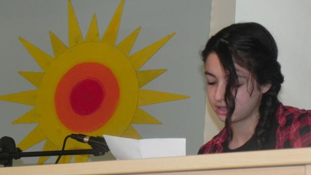 Ein junges Mädchen liest vor einem Mikrofon, im Hintergrund eine Sonne an der Wand.