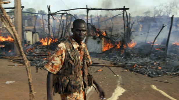 Ein Mann in Militärkleidung steht vor einem zerstörten und brennenden Dorf.