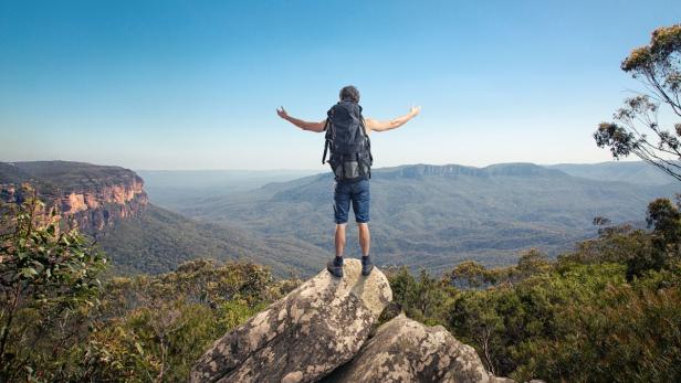 Ein Wanderer steht mit ausgebreiteten Armen auf einem Felsen und blickt auf die grüne Landschaft.