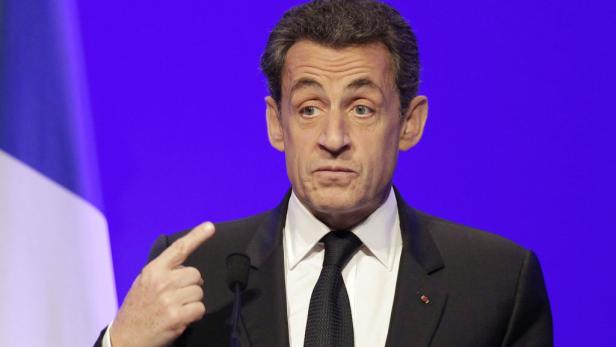 Nicolas Sarkozy gestikuliert vor einer blau-weißen Flagge.