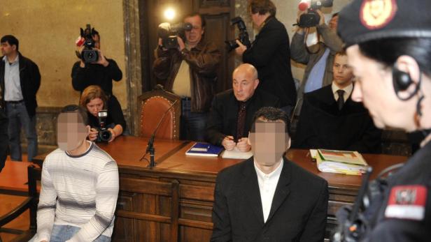 Zwei Angeklagte sitzen vor Gericht, während Fotografen und eine Polizistin anwesend sind.