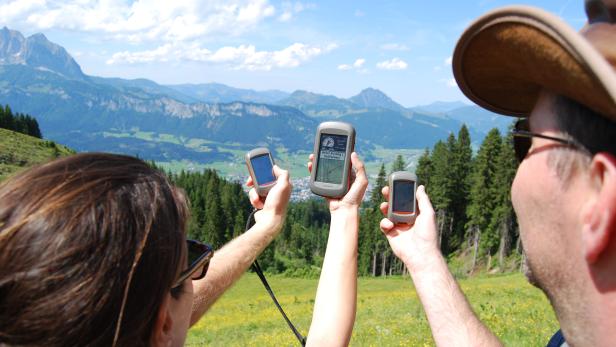 Zwei Personen halten GPS-Geräte vor einer Berglandschaft in die Höhe.