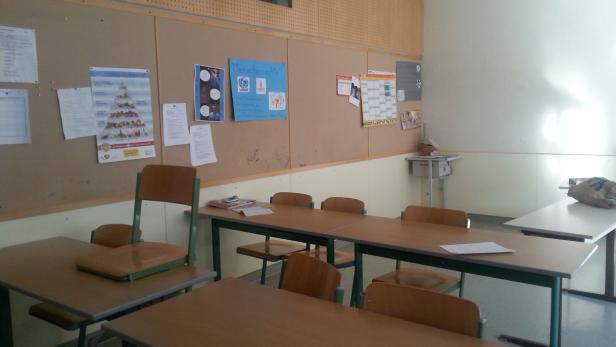 Ein Klassenzimmer mit Tischen, Stühlen und einer Pinnwand mit Postern.