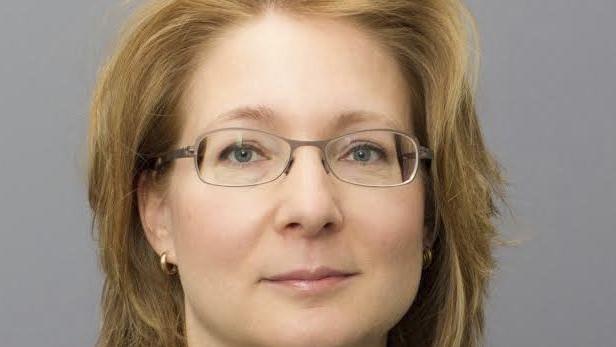 Porträt einer Frau mit blonden Haaren und Brille vor einem grauen Hintergrund.