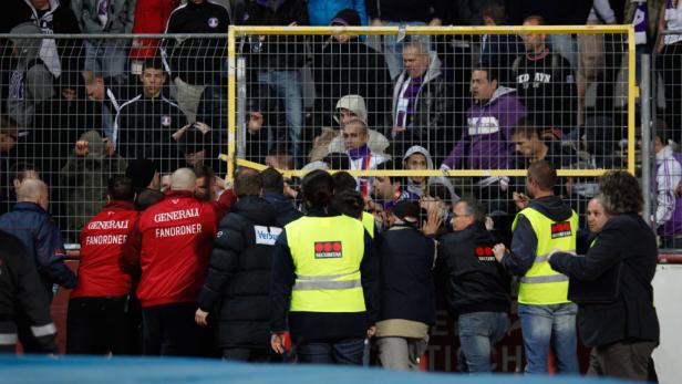 Ordner und Fans stehen an einem Gitter bei einem Fußballspiel.