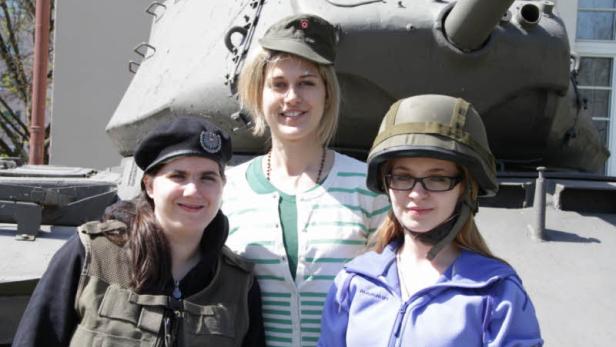 Drei Frauen posieren vor einem Panzer, zwei davon tragen militärische Kopfbedeckung.