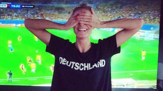 Eine Frau mit einem T-Shirt mit der Aufschrift „Deutschland“ hält sich die Augen zu und lächelt vor einem Fußballspiel im Fernsehen.