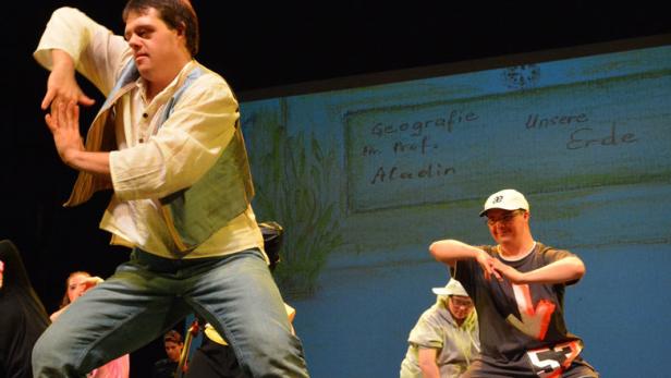 Eine Gruppe von Menschen mit Behinderung tanzt auf einer Bühne vor einer Leinwand mit dem Titel „Geografie“.