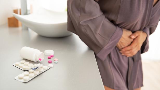 Pillen, die wie Antibiotika aussehen auf einem Tisch, eine Frau hält sich im Hintergrund den Bauch