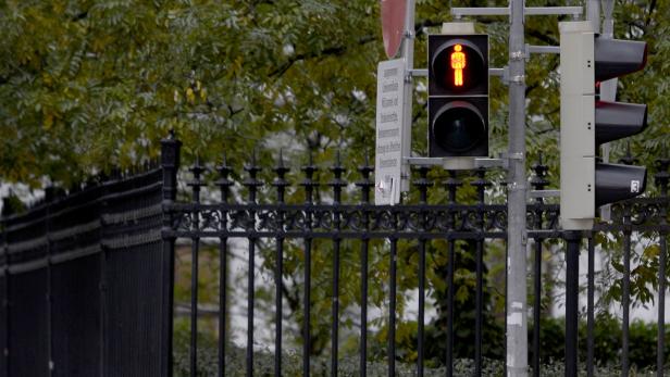 Eine rote Fußgängerampel vor einem schwarzen Zaun und Bäumen.