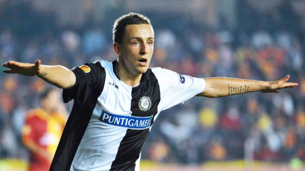 Ein Fußballspieler in einem schwarz-weiß gestreiften Trikot jubelt mit ausgestreckten Armen.