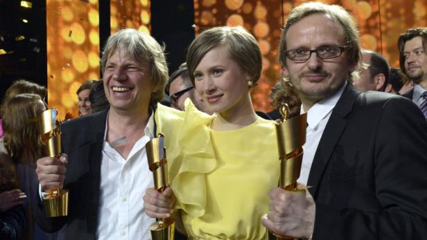 Drei Personen halten goldene Auszeichnungen vor einem festlichen Hintergrund.