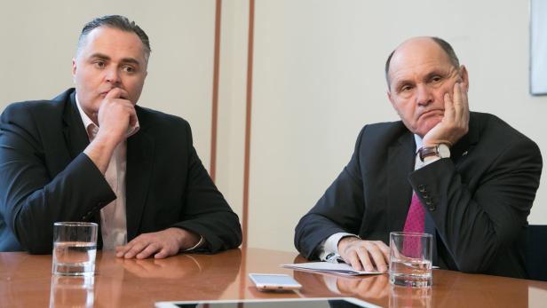 Zwei Männer in Anzügen sitzen an einem Tisch mit Gläsern Wasser und elektronischen Geräten.